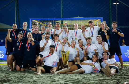 A beach soccer team poses triumphantly on the sand with their medals and trophy, celebrating a victory under stadium lights.