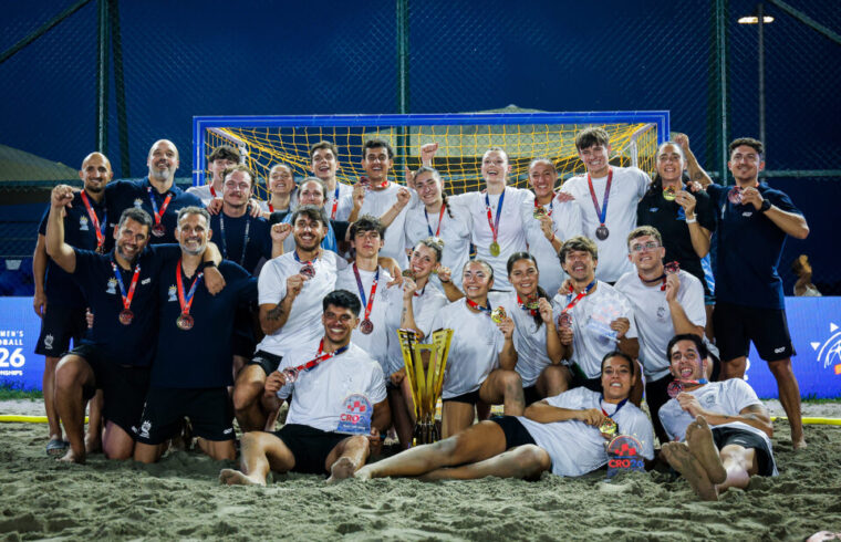 A beach soccer team poses triumphantly on the sand with their medals and trophy, celebrating a victory under stadium lights.